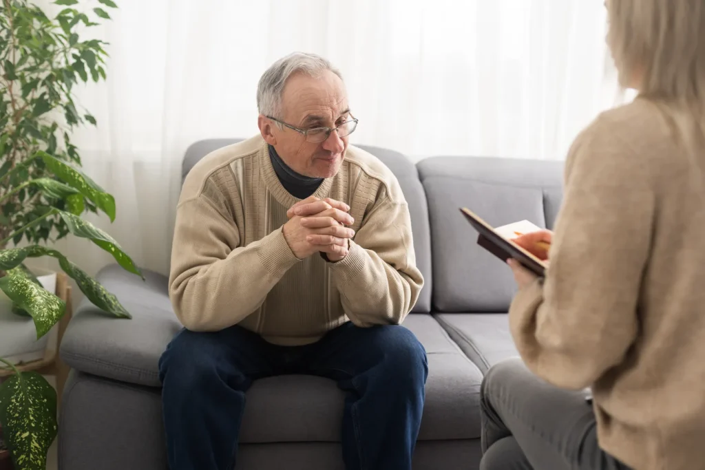 An older gentleman sits on a couch while someone talks to him holding paperwork.