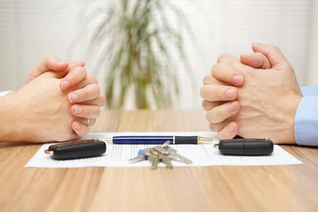 Two people sitting at a desk with their hands folded and in between them are keys, a contract and a pen.