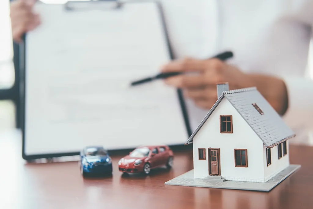 A miniture house with two toy cars on a desk, in the backgroiund is someone holding up a clipboard and pointing with a pen where to sign.