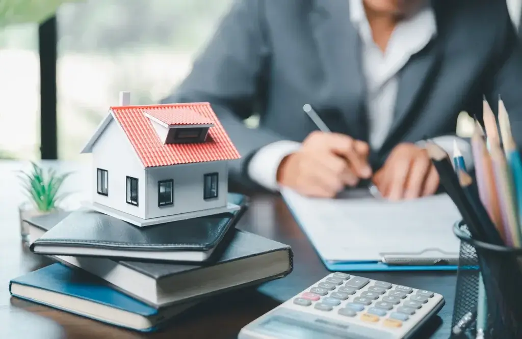 Person signing documents behind a miniature house on notebooks, representing the need to update an estate plan after inheriting property in Texas.